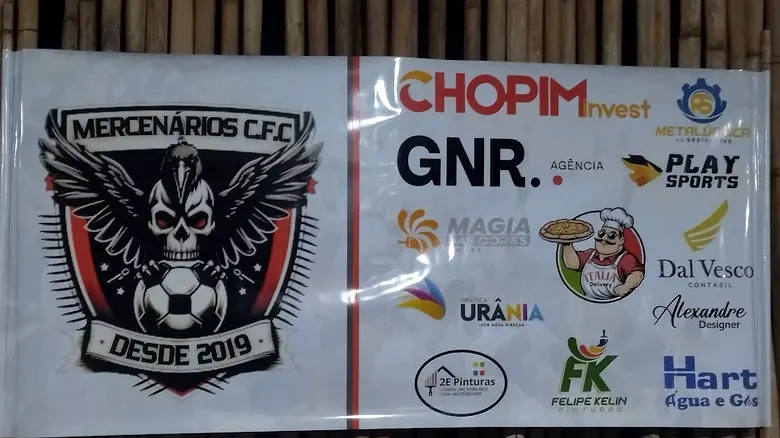Banner em lona do time Mercenários C.F.C. com os logos dos patrocinadores, instalado em cerca